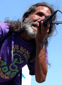 A brown skinned man with a short gray beard and long black and gray hair leans forward speaking into a microphone he's holding. He is bent forward and wearing a purple ADAPT T-shirt. He looks very intense